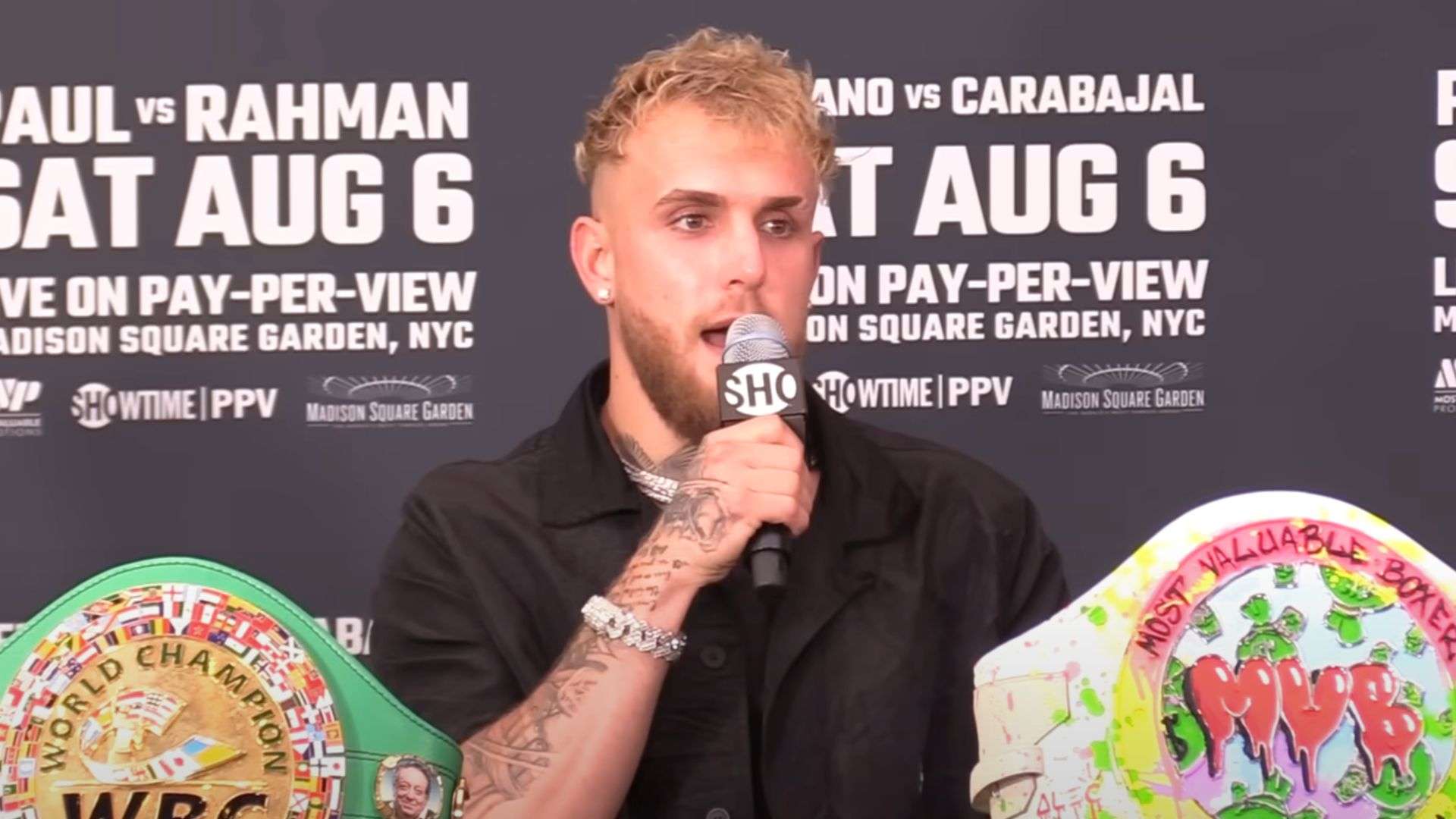 Jake Paul holding mic at press conference surronded by boxing belts