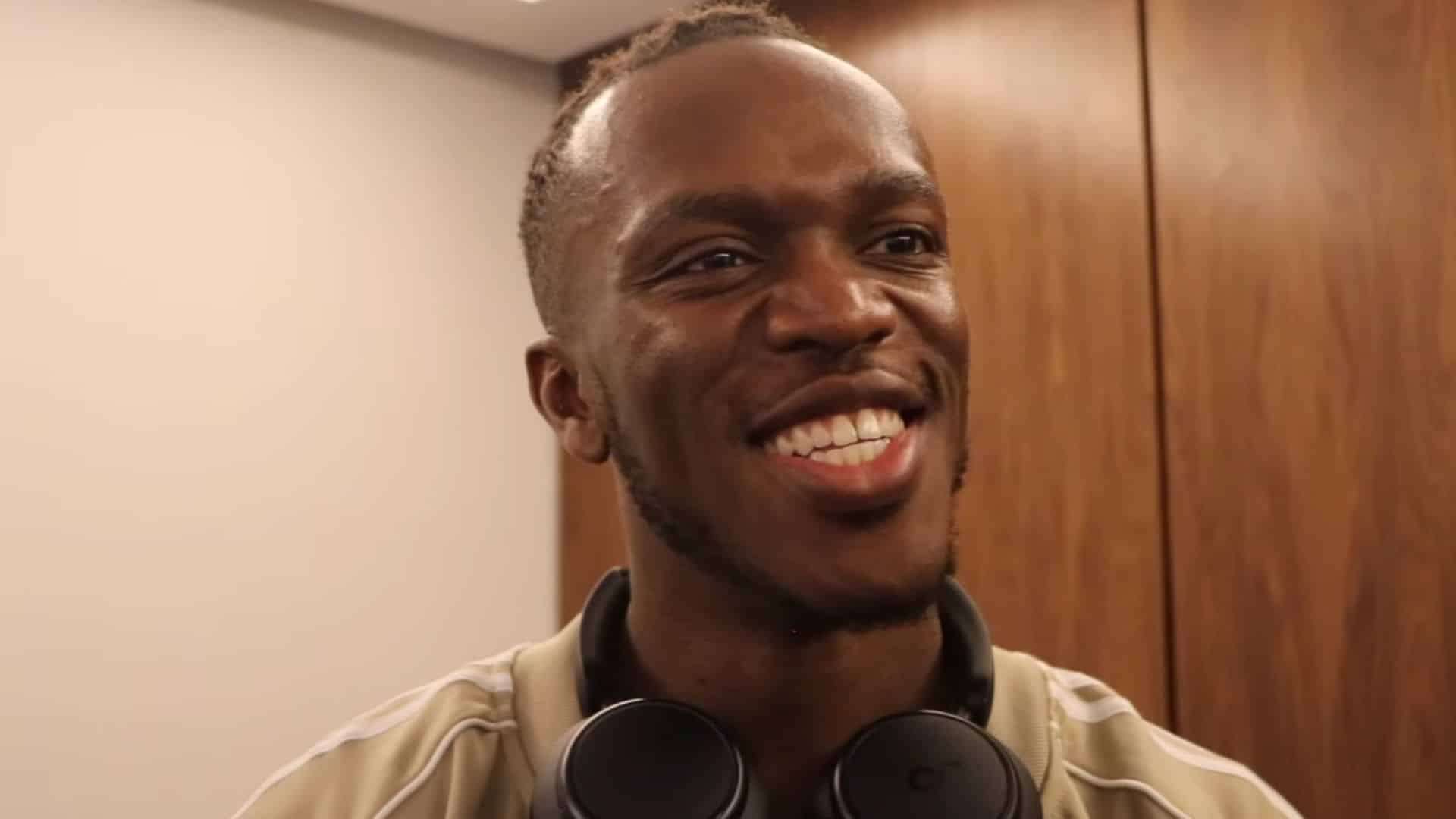 KSI smiling at camera in brown shirt and with headphones on