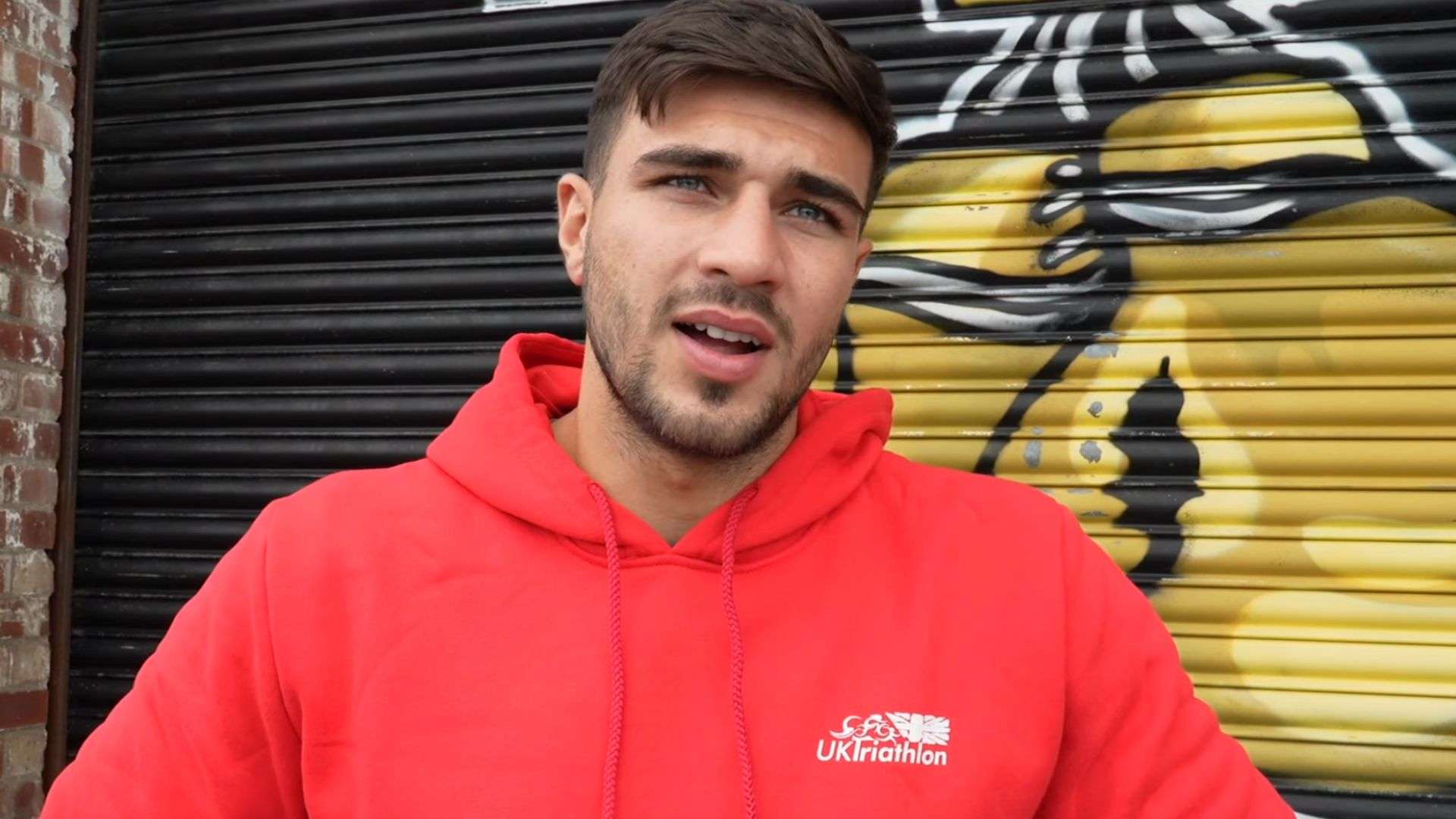 Tommy Fury in red hoodie talking to camera in front of gold boxing gloves painted on garage door