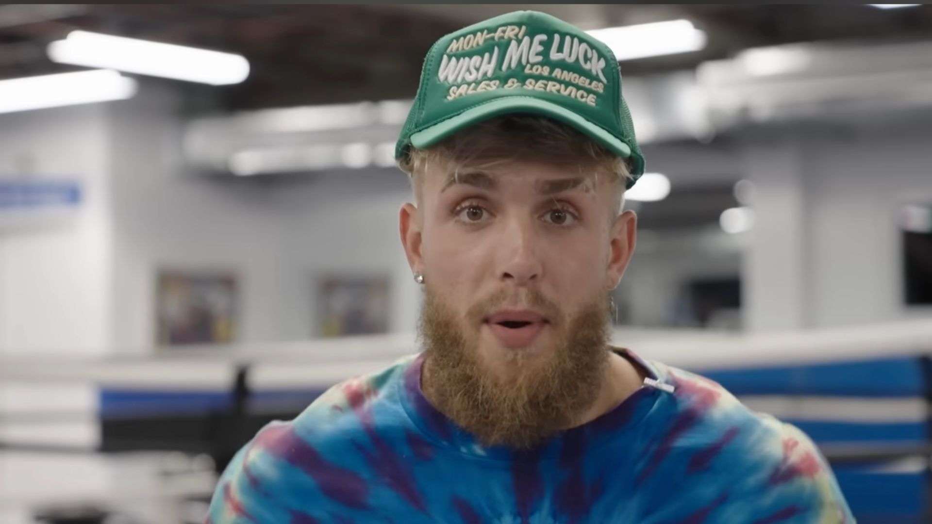 Jake Paul in blue and purple shirt wearing green hat in front of boxing ring