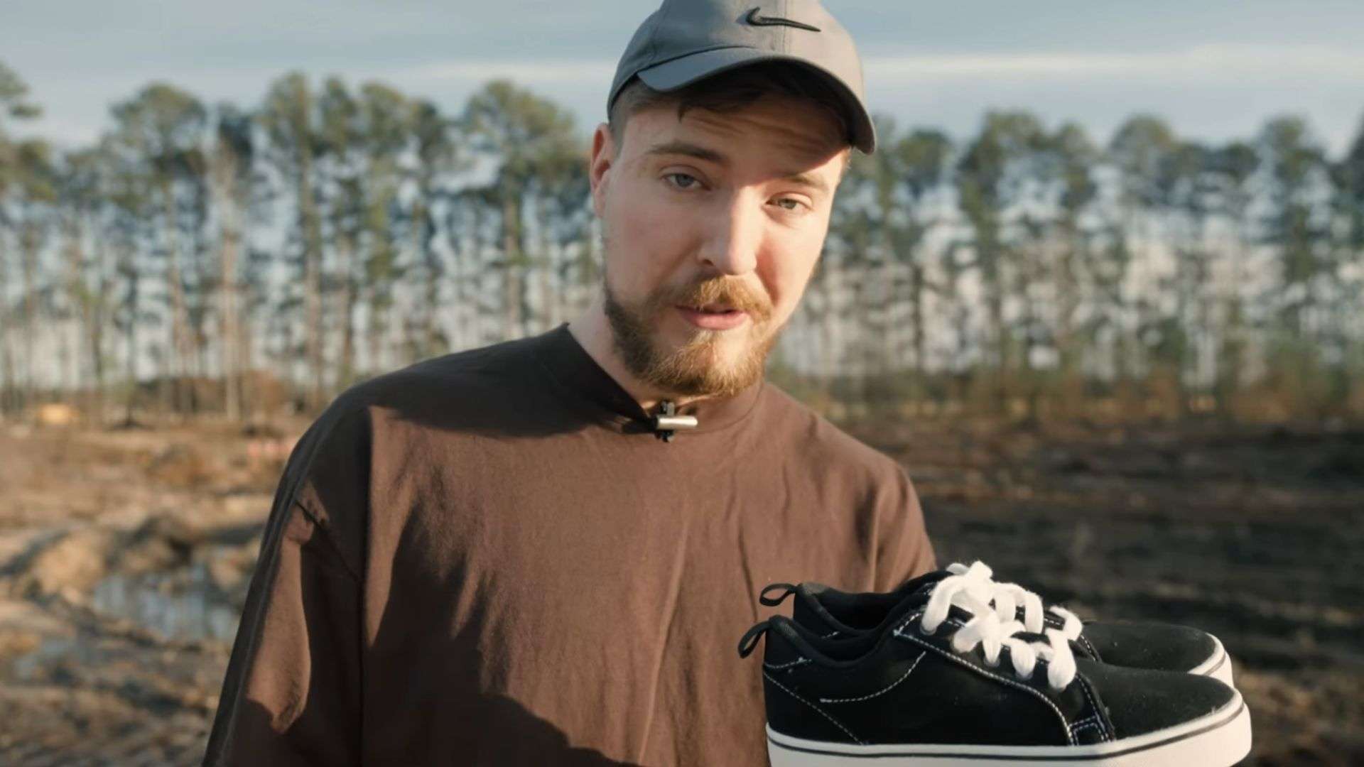 MrBeast in black shirt and gray hat holding black pair of shoes and talking to camera