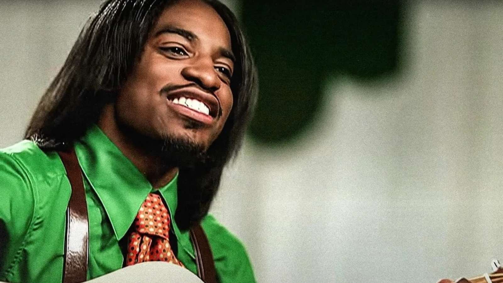 André 3000 in a green button up shirt and suspenders playing a guitar