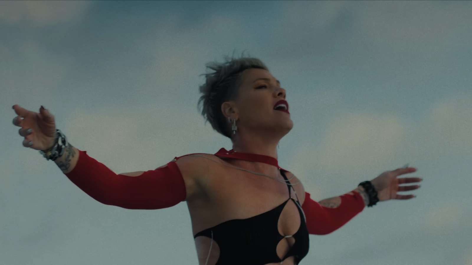 Singer P!nk with her arms open wide in front of a cloudy blue sky