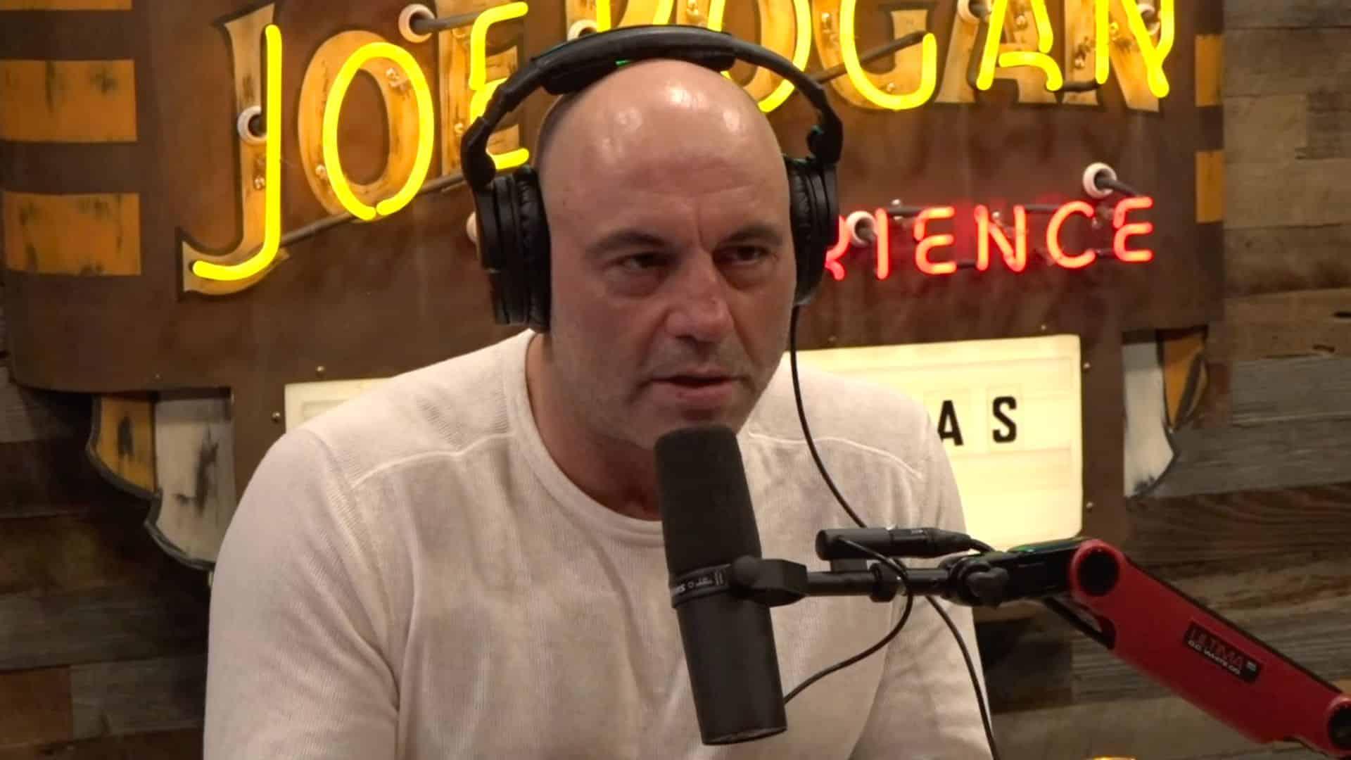 Joe Rogan talking to mic on podcast set with white shirt on