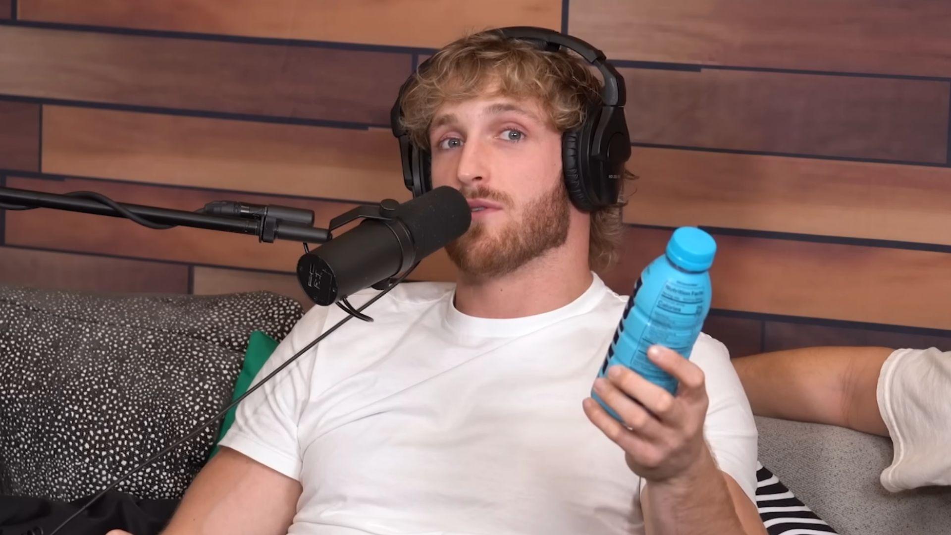 Logan Paul in white t-shirt holding blue bottle of prime drink next to microphone