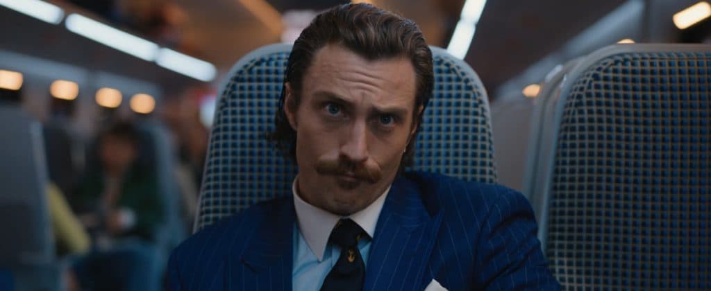 Aaron Taylor-Johnson in Bullet Train. He sits on a train chair, looking irritated.