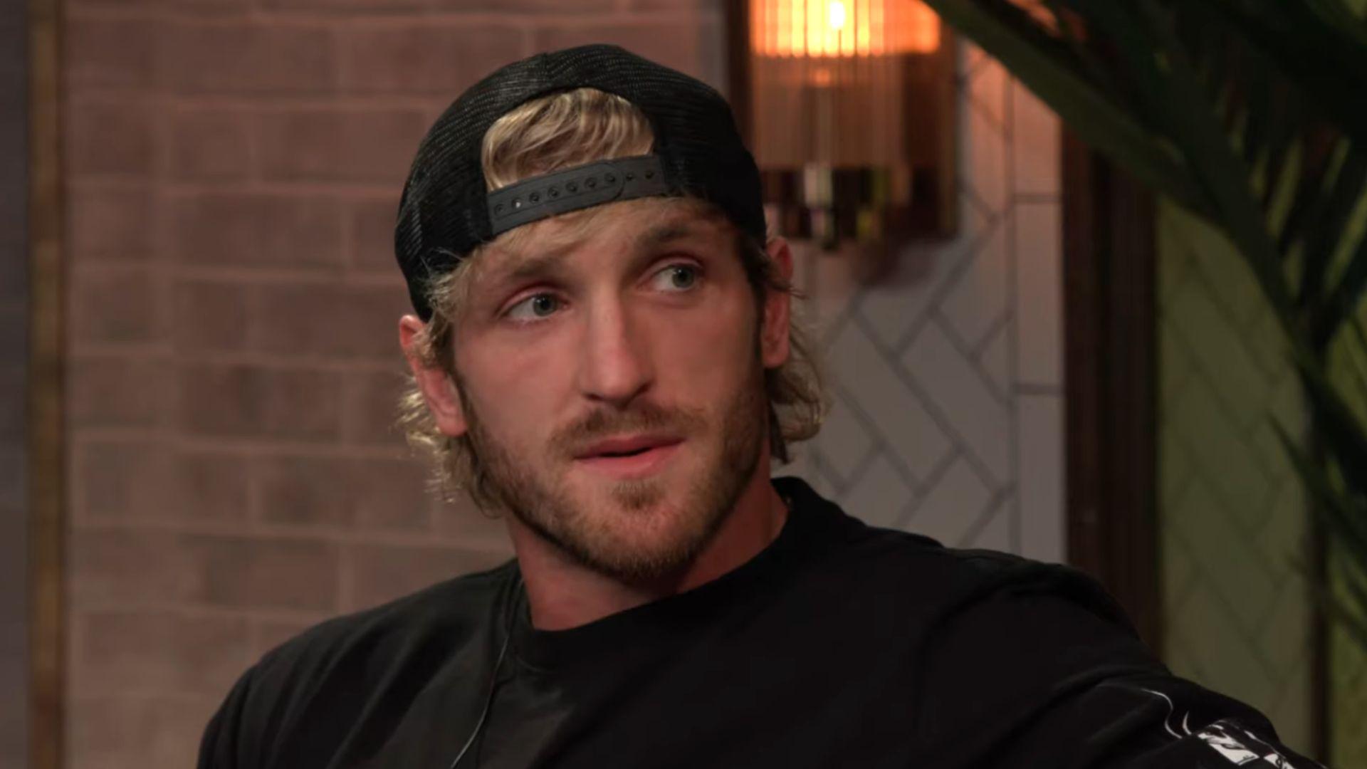 Logan Paul in black hat and shirt talking to camera on podcast couch
