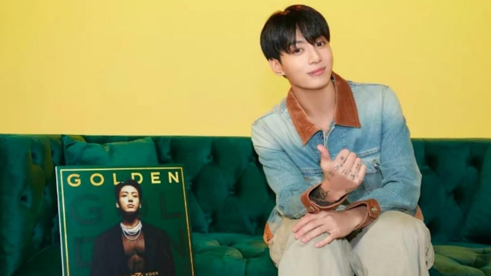 Jungkook posing with his thumb up and seated on a green sofa with his album, Golden.