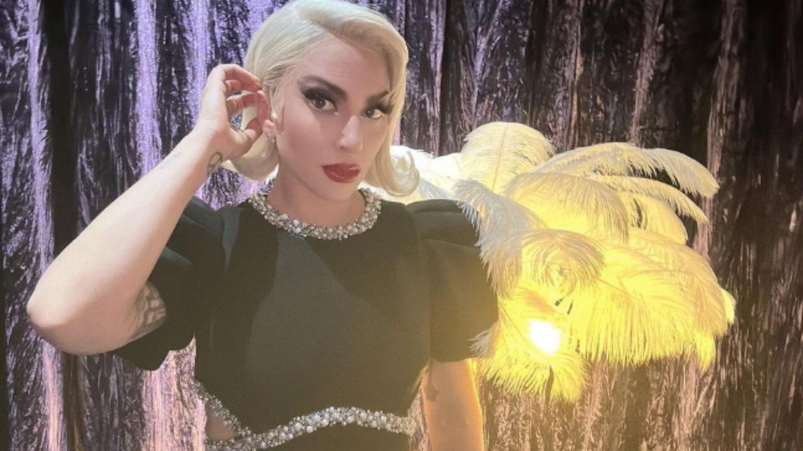 Lady Gaga posing in a black dress with diamonds in front of a feathered light.