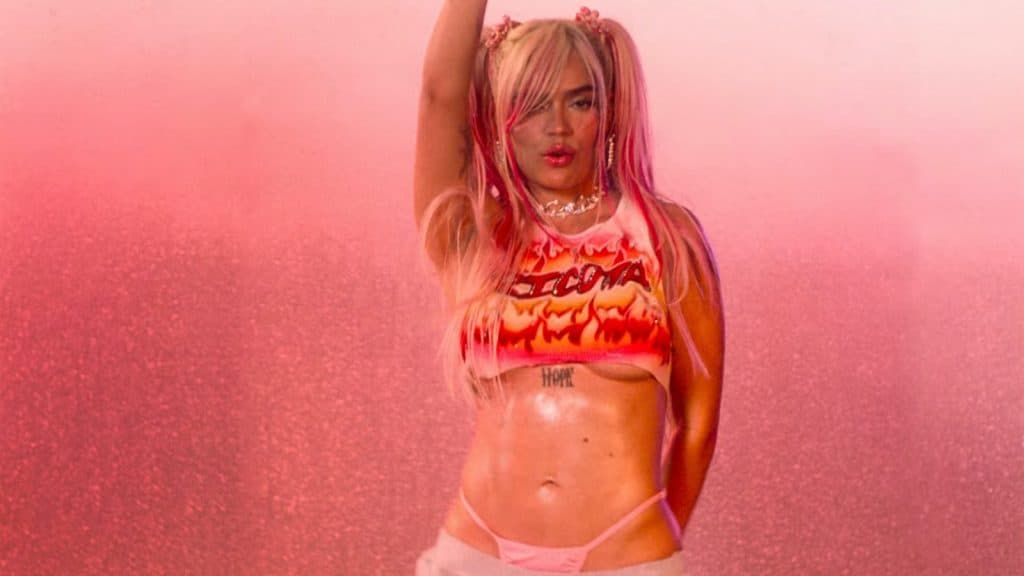 Karol G performin gin front of a pink backdrop with her right hand in the air