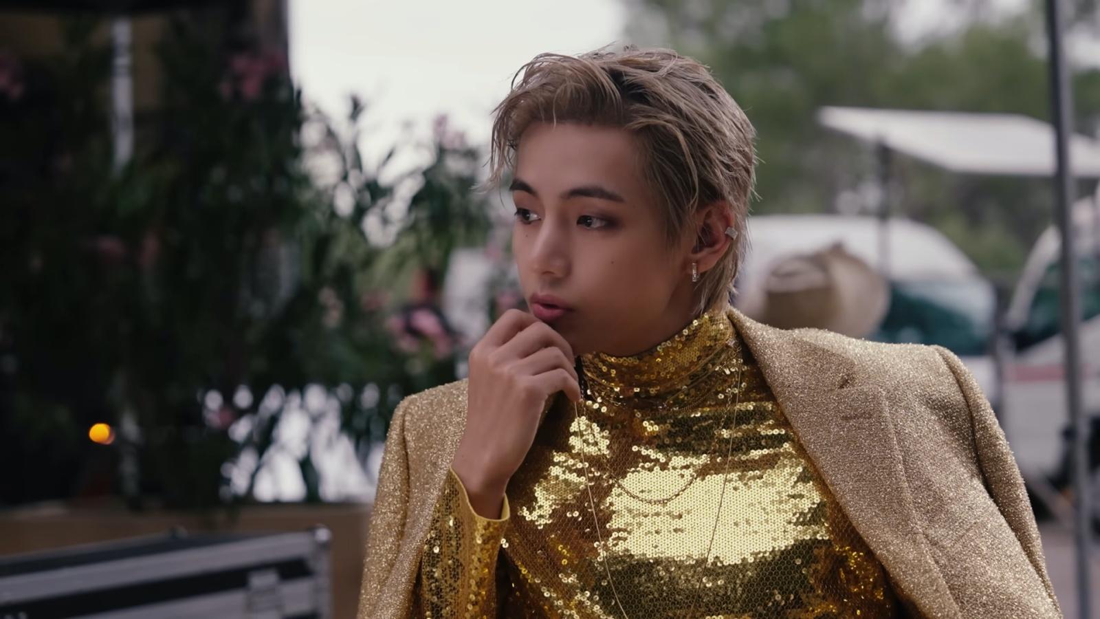 BTS' V sitting in a chair and wearing a gold outfit