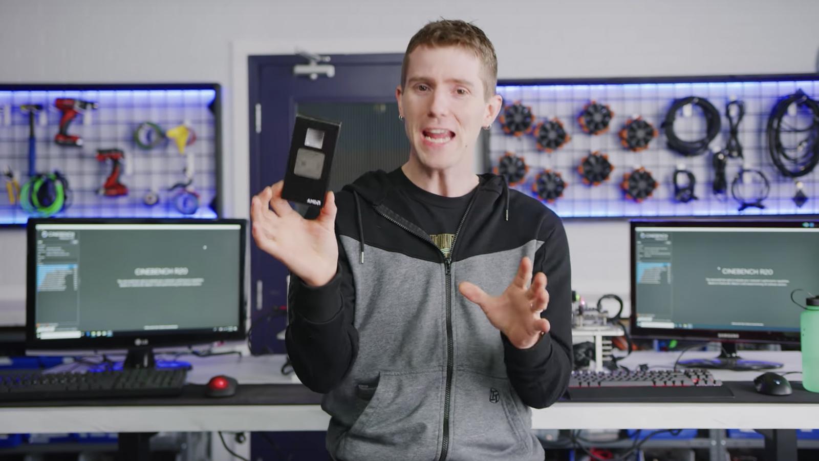 Linus holds a GPU in his hand in office with two monitors behind him