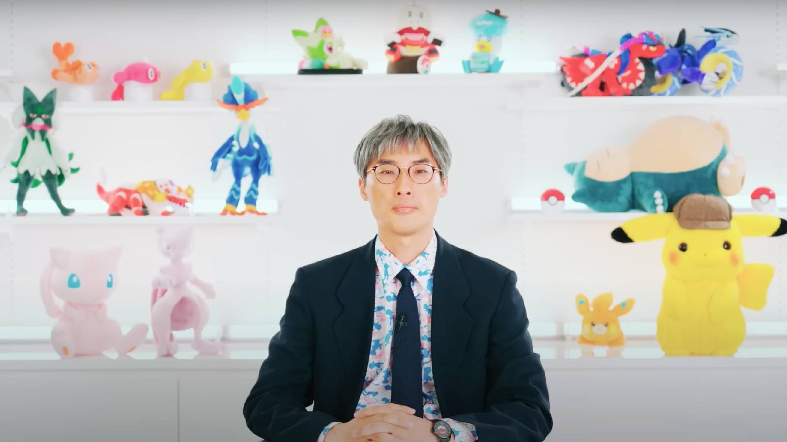 The Pokemon Company COO Takato Utsunomiya sits in a white room with a background filled with various POkemon plush. Dressed in a dark suit, a colourful shirt, and with think round glasses, Takato is sitting down and facing forward to present a Pokemon Presents video presentation