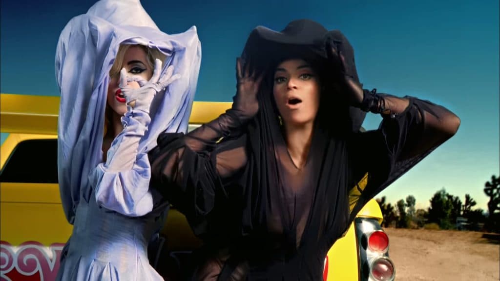 Beyoncé & Lady Gaga wearing lacy costumes and standing in the desert