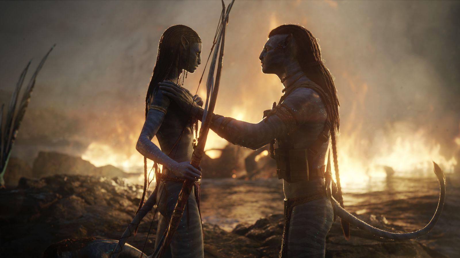 Jake Sully and Neytiri in Avatar 2. They stand with burning flames in the background.