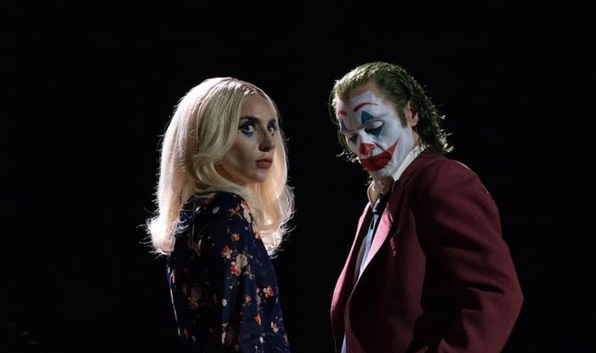 Joaquin Phoenix as Arthur Fleck and Lady Gaga as Harley Quinn in Joker 2. They stand together in pitch bdarkness, Arthur dressed as his Joker persona.