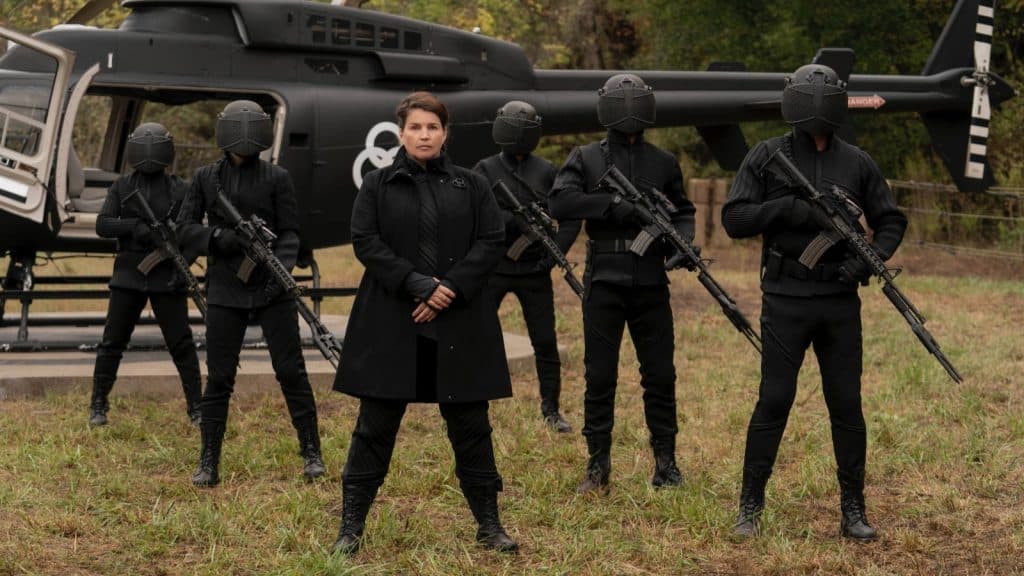 The CRM in The Walking Dead standing dressed in black in front of a helicopter, holding guns