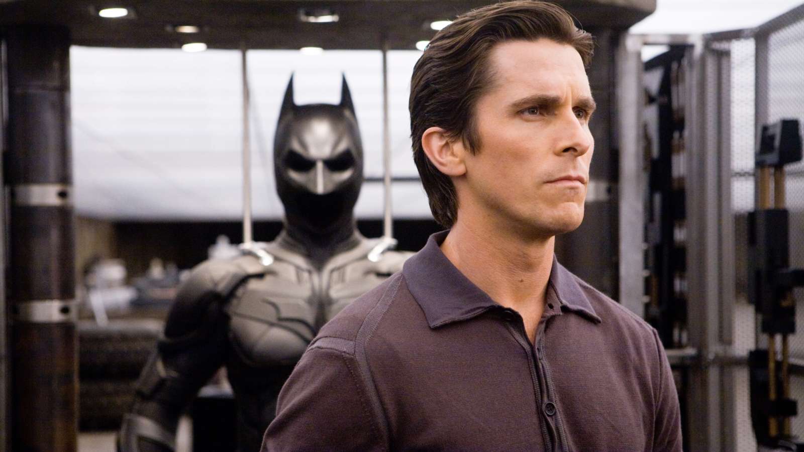 Christian Bale as Bruce Wayne in The Dark Knight, standing in front of the Batman suit