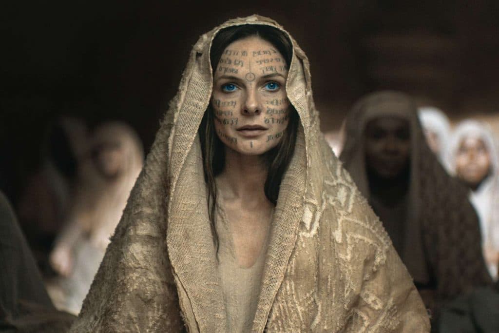 Rebecca Ferguson as Lady Jessica in Dune Part 2. Wearing a cloak and with scripture painted across, she gives a cold stare towards the viewer.