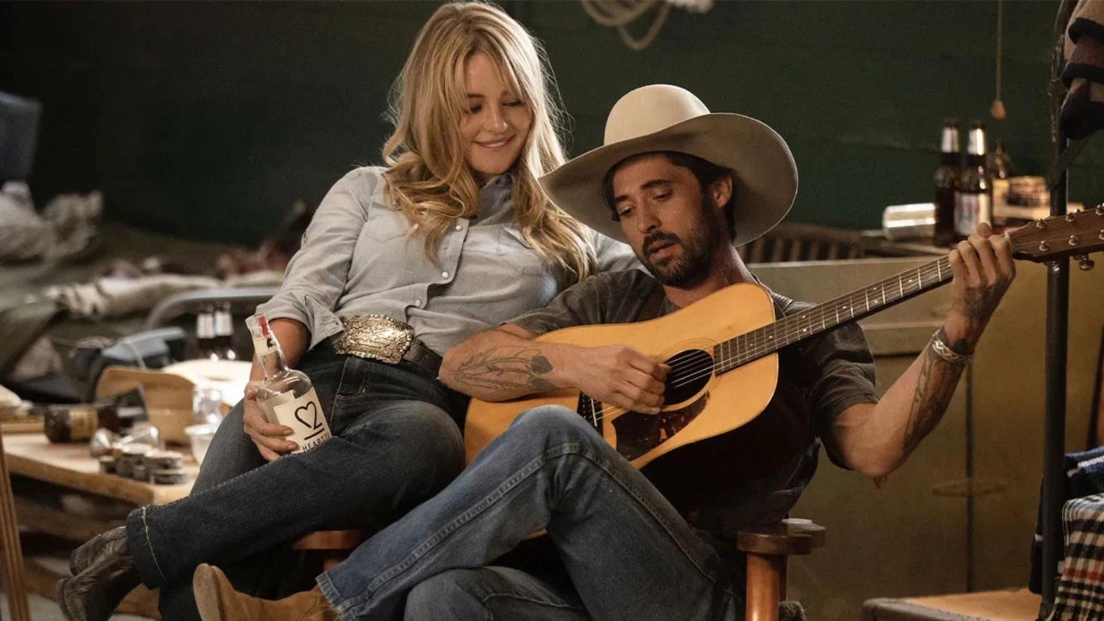 Hassie Harrison as Laramie and Ryan Bingham as Walker on Yellowstone, sitting on a chair with Walker holding a guitar