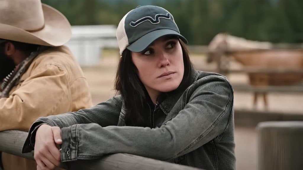 Eden Brolin as Mia on Yellowstone, wearing a baseball cap and leaning against a fence
