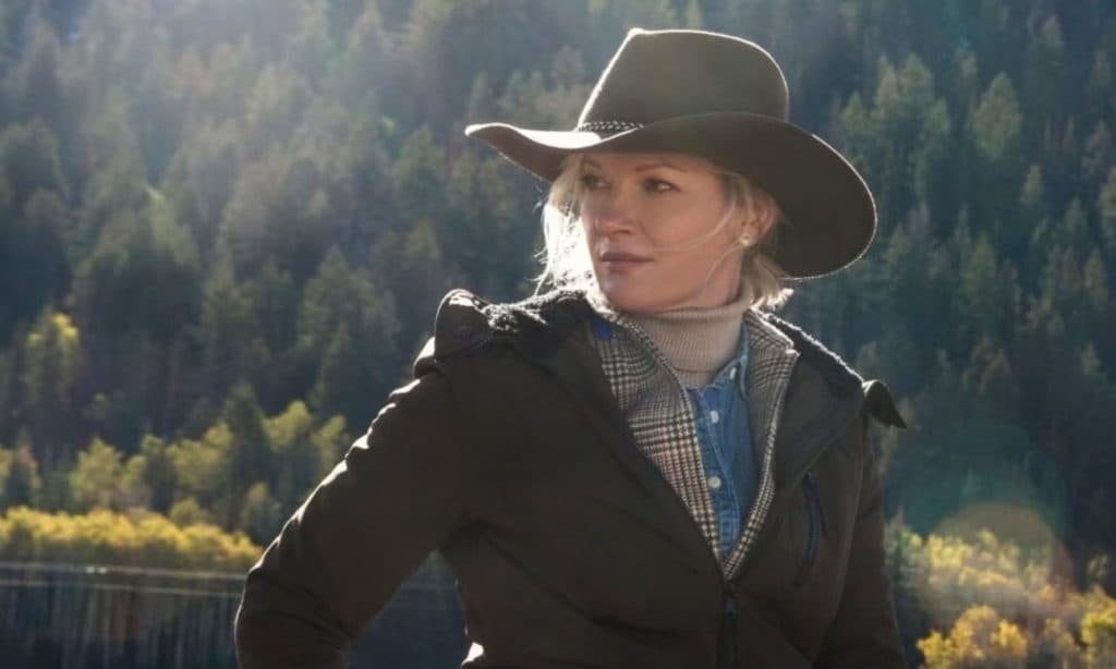 John Dutton's wife, Evelyn, standing against the woods and wearing a cowboy hat