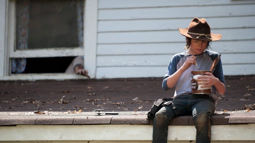 Chandler Riggs as Carl in The Walking Dead, sitting on a rooftop eating pudding