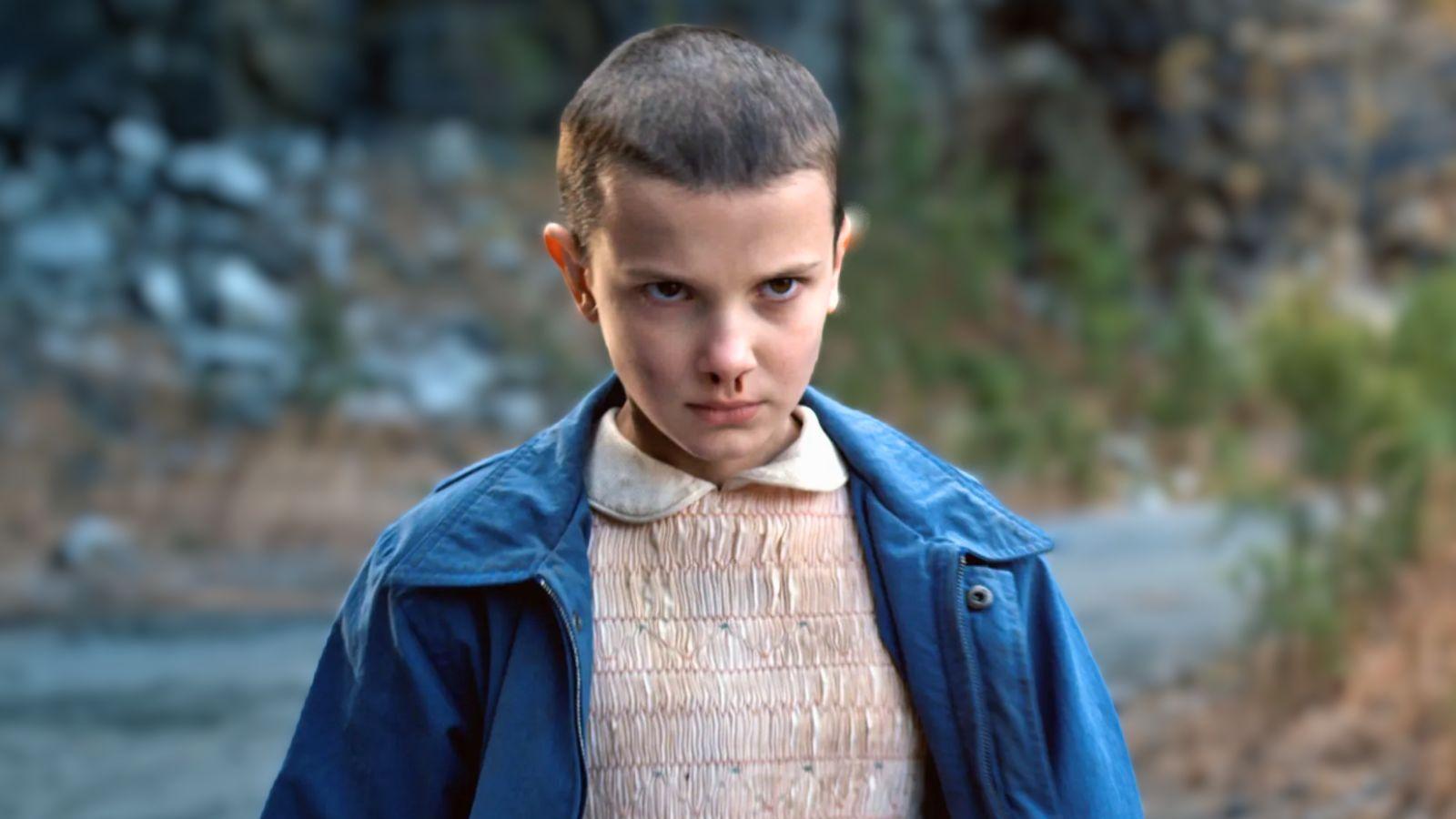 Millie Bobby Brown as Eleven in Stranger Things Season 1. She stands in woodland area, staring intensely with a nose bleed while preparing to use her powers.