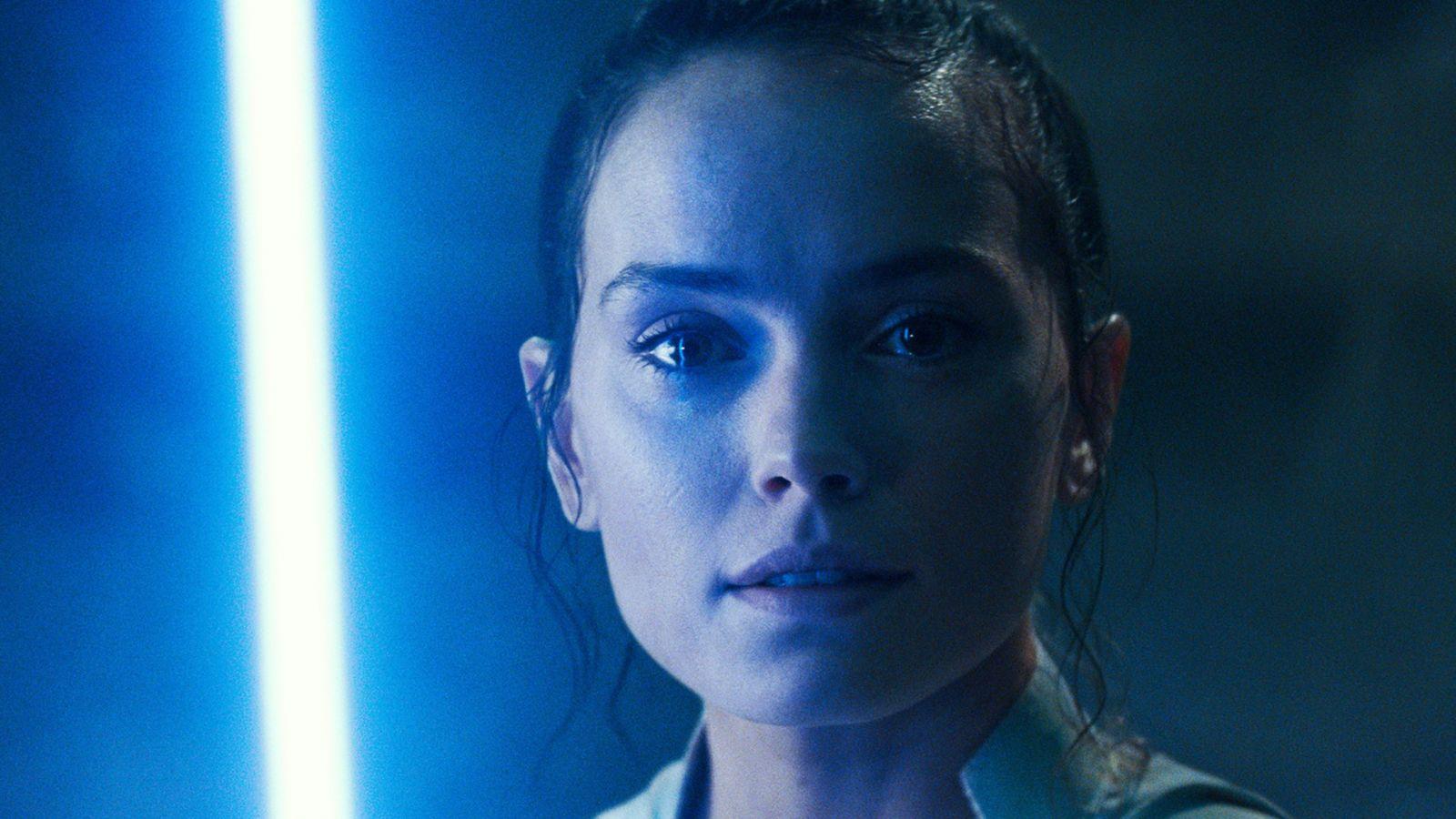 Daisy Ridley as Rey Skywalker in Star Wars The Force Awakens. The left side of her face is lit by blue lightsaber light, and she has tears in her eyes.