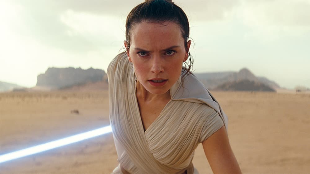 Daisy Ridley as Rey Skywalker in Star Wars The Rise of Skywalker. She is standing defensively with her blue lightsaber drawn, anticipating an attack and steadying herself.