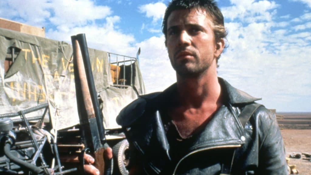 Mad Max in front of a rig holding a shotgun.