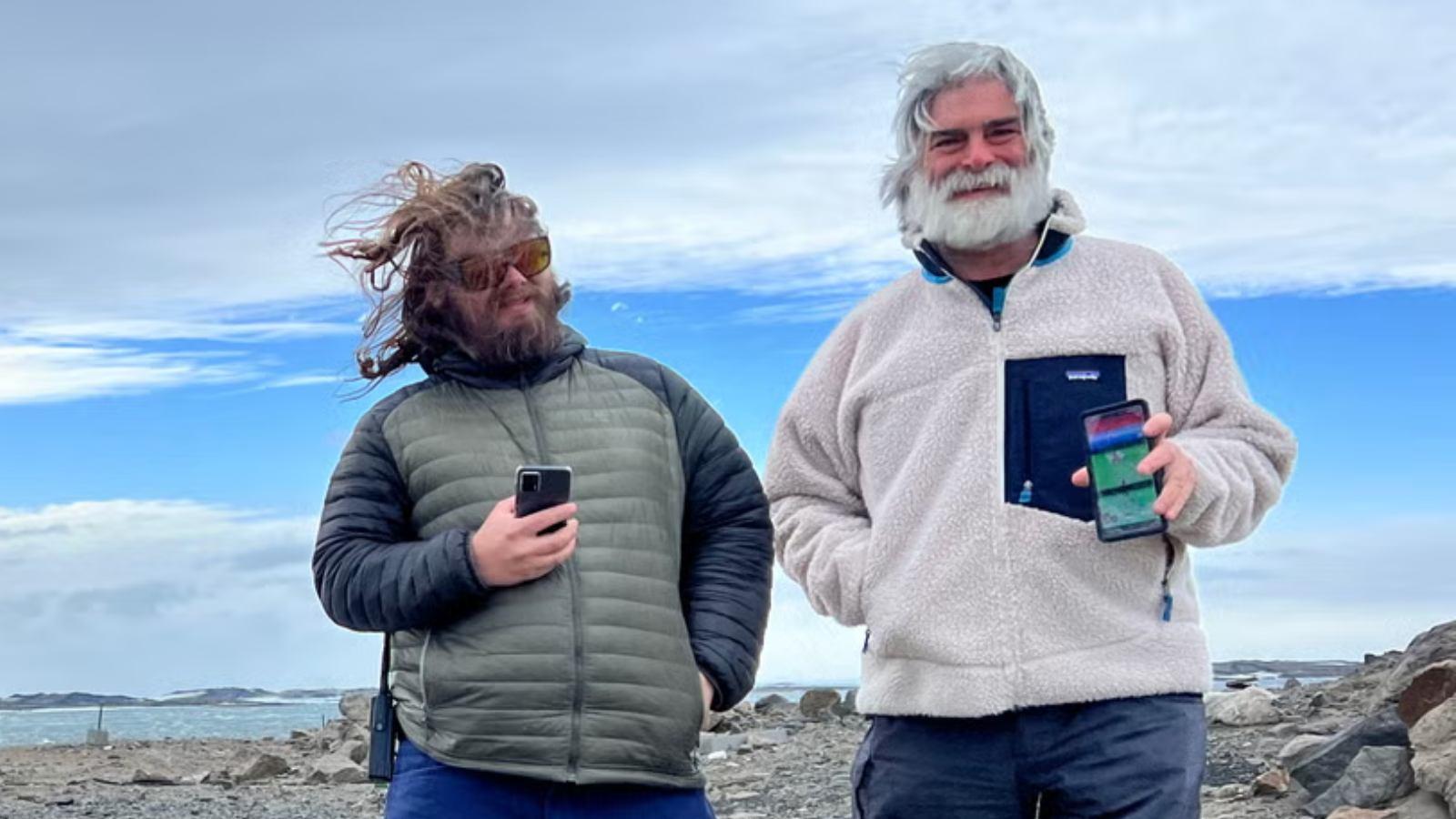 Two scientists stood in Antarctica hold their phones aloft, with Pokemon Go visible on the screens