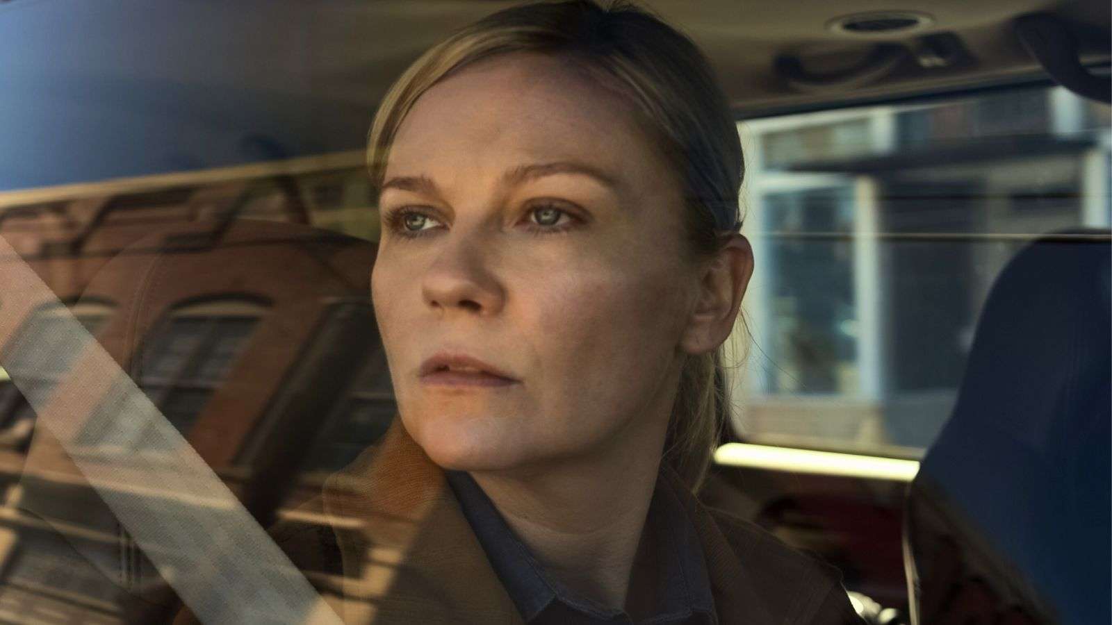 Kirsten Dunst in Civil War rides in the passenger seat of a car while looking out the window at buildings.