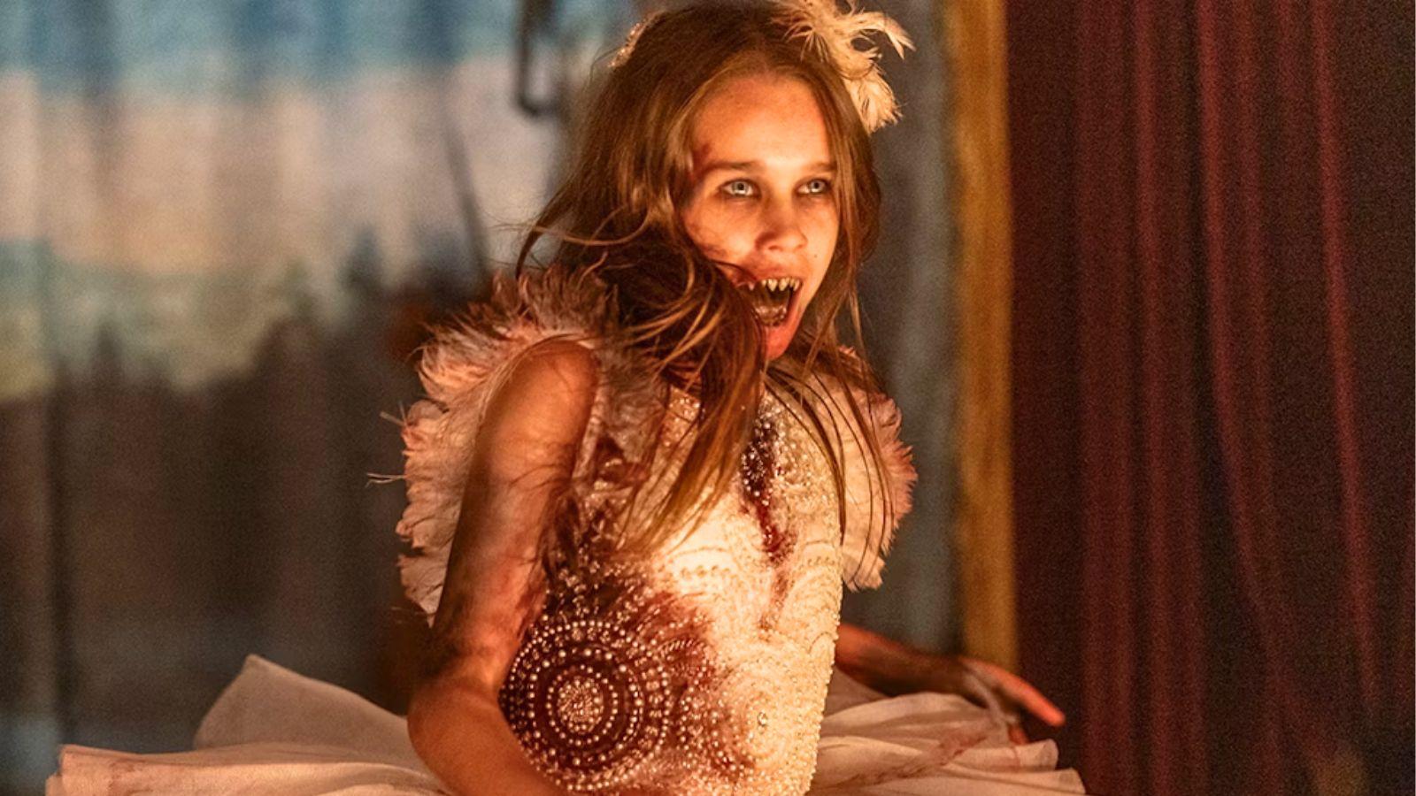 Alisha Weir as Abigail. She stands in a ballerina dress with blood stains and her fangs visible.